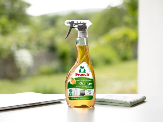 A Frosch multi-surface cleaner orange trigger bottle stands next to a cleaning cloth and laptop on a table